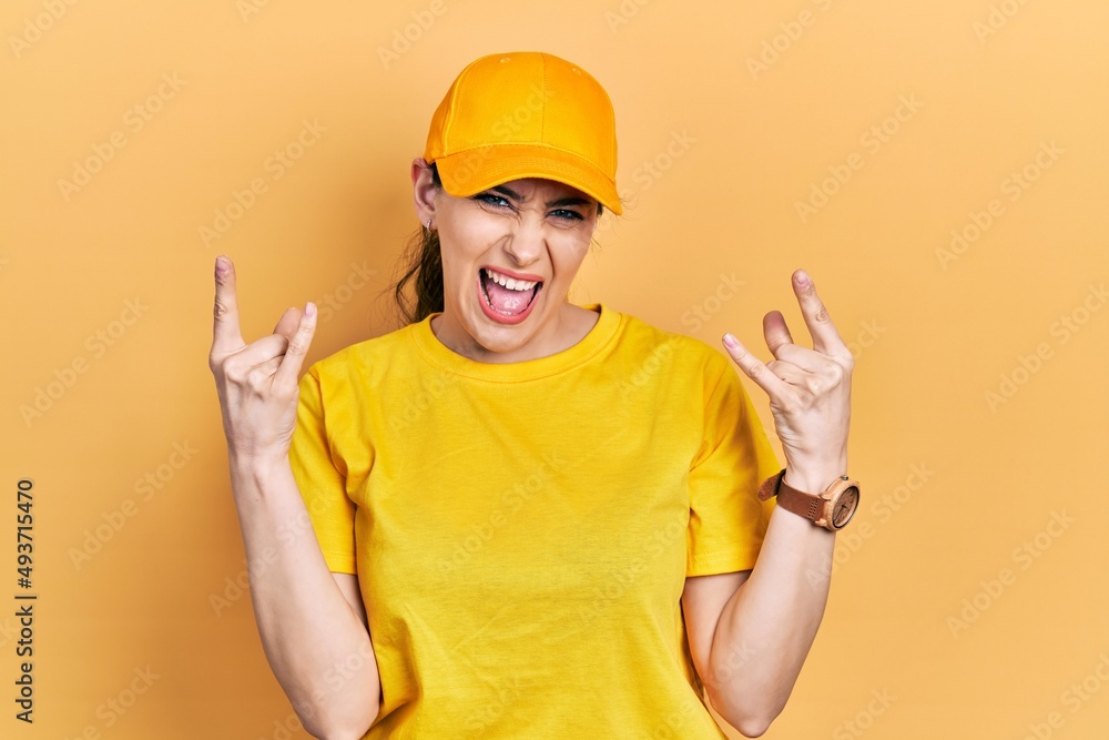 Young hispanic woman wearing delivery uniform and cap shouting with crazy expression doing rock symbol with hands up. music star. heavy concept.