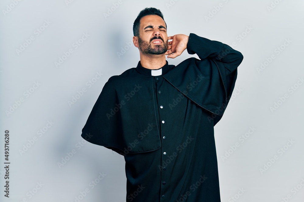 Handsome hispanic man with beard wearing catholic priest robe suffering of neck ache injury, touching neck with hand, muscular pain Handsome hispanic man with beard wearing catholic priest robe suffering of neck ache injury, touching neck with hand, muscular pain