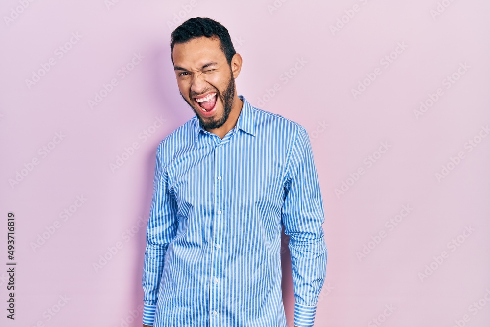 Hispanic man with beard wearing casual blue shirt winking looking at the camera with sexy expression, cheerful and happy face. Hispanic man with beard wearing casual blue shirt winking looking at the camera with sexy expression, cheerful and happy face.