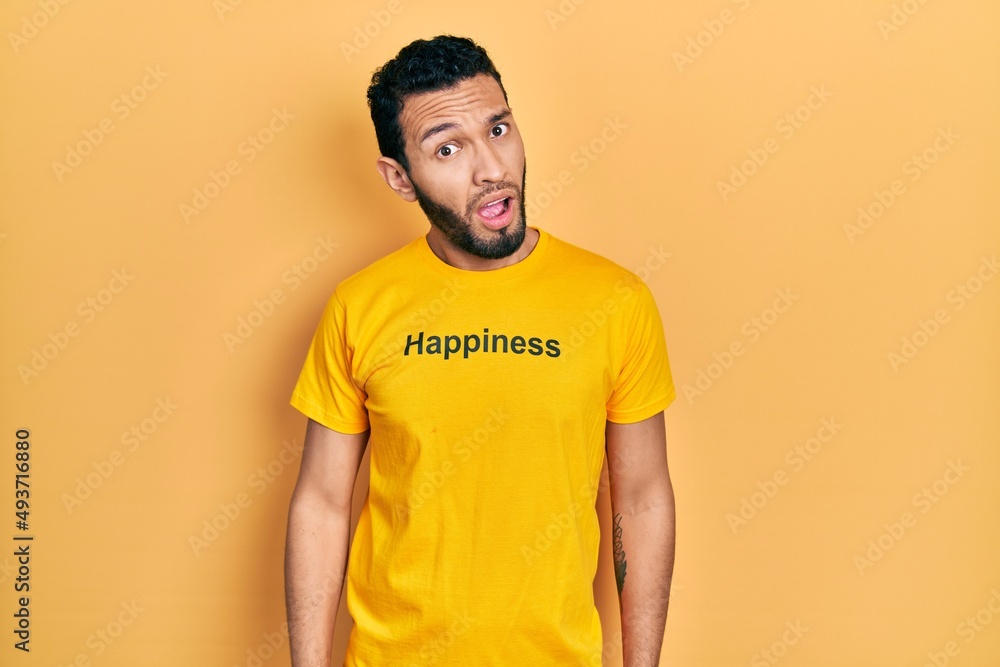 Hispanic man with beard wearing t shirt with happiness word message in shock face, looking skeptical and sarcastic, surprised with open mouth Hispanic man with beard wearing t shirt with happiness word message in shock face, looking skeptical and sarcastic, surprised with open mouth
