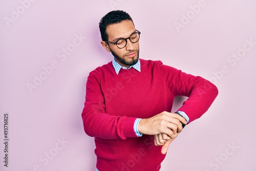 Hispanic man with beard wearing business shirt and glasses checking the time on wrist watch, relaxed and confident