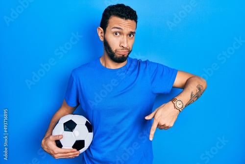 Hispanic man with beard holding soccer ball pointing down looking sad and upset, indicating direction with fingers, unhappy and depressed.