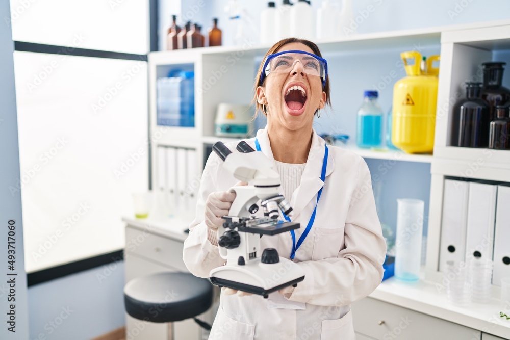Young brunette woman working at scientist laboratory with microscope angry and mad screaming frustrated and furious, shouting with anger looking up. Young brunette woman working at scientist laboratory with microscope angry and mad screaming frustrated and furious, shouting with anger looking up.