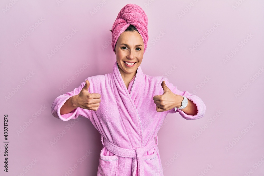Young blonde woman wearing shower towel cap and bathrobe success sign doing positive gesture with hand, thumbs up smiling and happy. cheerful expression and winner gesture. Young blonde woman wearing shower towel cap and bathrobe success sign doing positive gesture with hand, thumbs up smiling and happy. cheerful expression and winner gesture.