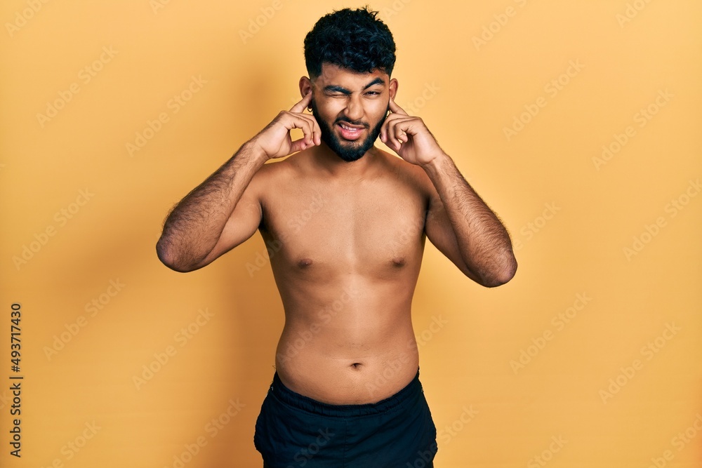 Arab man with beard wearing swimwear shirtless covering ears with fingers with annoyed expression for the noise of loud music. deaf concept. Arab man with beard wearing swimwear shirtless covering ears with fingers with annoyed expression for the noise of loud music. deaf concept.