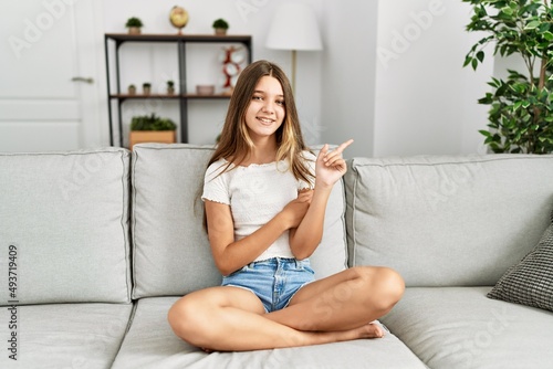 Young brunette teenager sitting on the sofa at home with a big smile on face, pointing with hand and finger to the side looking at the camera.