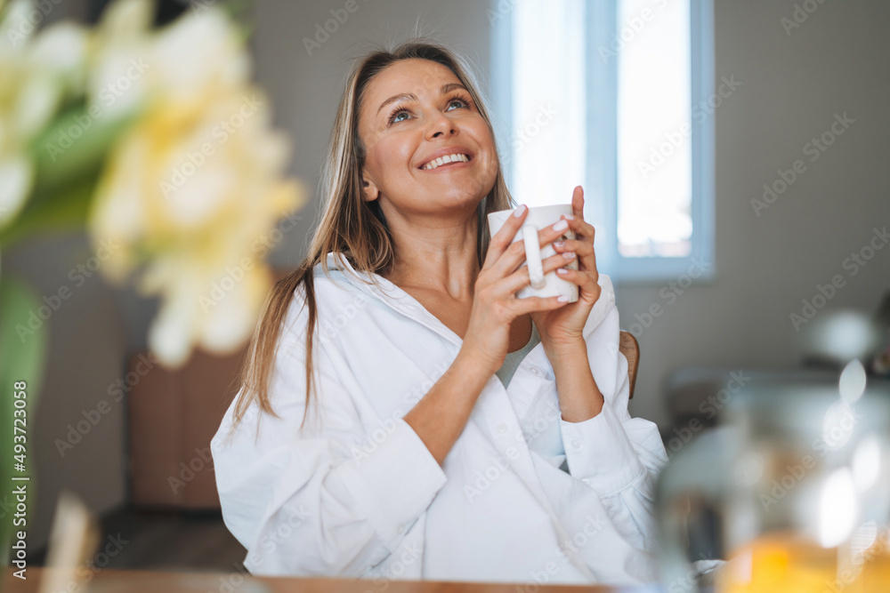 Young beautiful happy woman with blonde long hair in white shirt drinking tea with bouquet of yellow flowers in vase on dinner table in bright interior at home Young beautiful happy woman with blonde long hair in white shirt drinking tea with bouquet of yellow flowers in vase on dinner table in bright interior at home