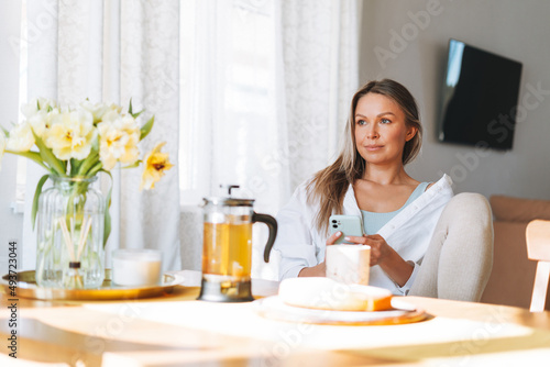 Young beautiful happy woman with blonde long hair in white shirt drinking tea using mobile phone at sunny living room at the home