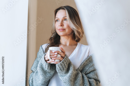 Young beautiful woman forty year with blonde long curly hair in cozy knitted grey sweater with cup of tea in hands in bright interior at the home
