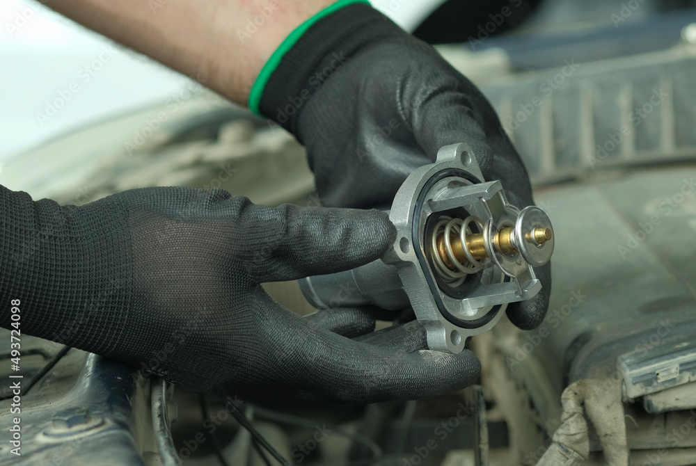 Repair and maintenance in a car service center. Spare parts. Thermostat for the engine. Close-up.  An auto mechanic holds a new thermostat in his hands against the background of the engine compartment