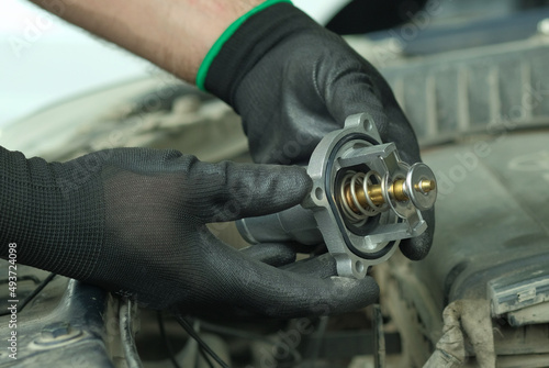 Repair and maintenance in a car service center. Spare parts. Thermostat for the engine. Close-up. An auto mechanic holds a new thermostat in his hands against the background of the engine compartment