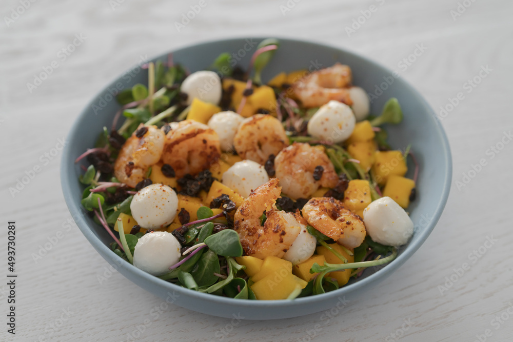 Salad with mango, mozzarella, shrimps and mixed microgreens in blue bowl on white oak table Salad with mango, mozzarella, shrimps and mixed microgreens in blue bowl on white oak table