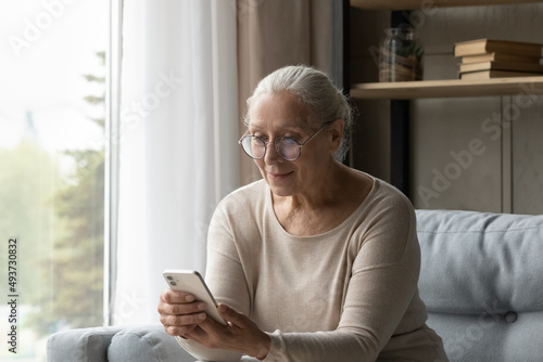 Aged woman in glasses holding smartphone seated on sofa at home, use mobile application, read social media news. Older generation use modern tech, easy comfort remote electronic services usage concept