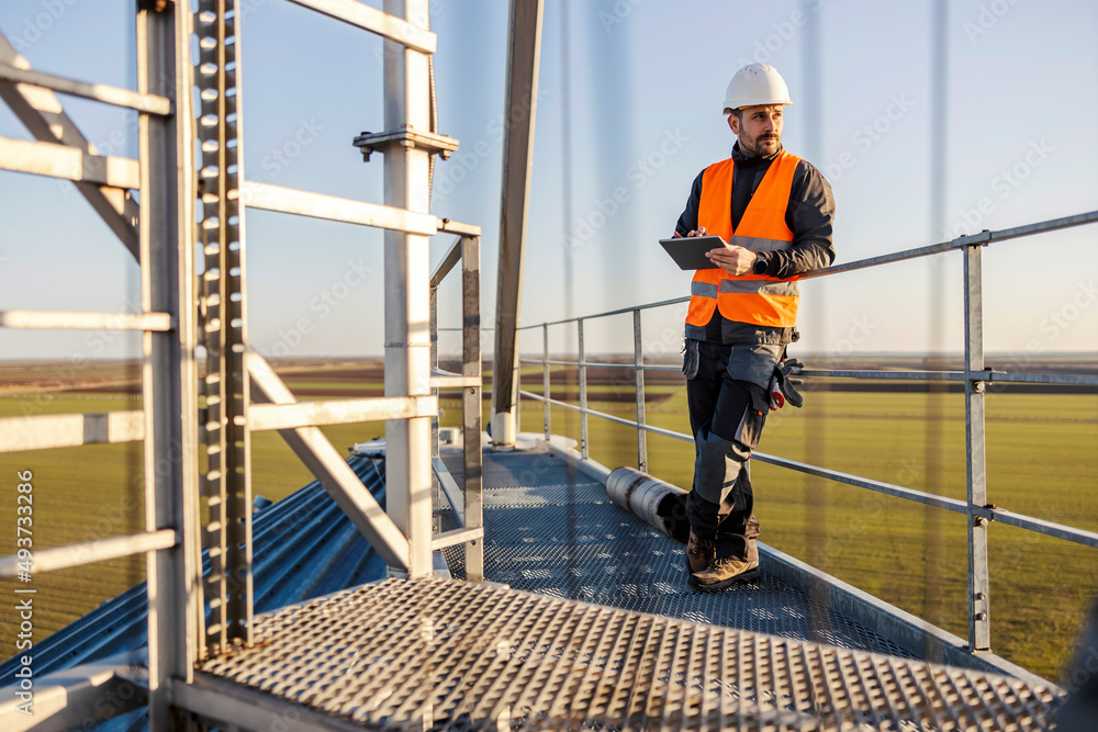 An industry worker standing on top of the silo and scrolling on tablet. An industry worker standing on top of the silo and scrolling on tablet.