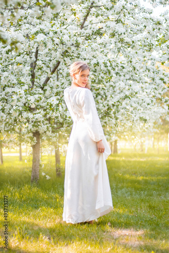 Young woman in a white dress against the backdrop of blooming apple trees. The girl poses against the background of flowers in the spring park. 