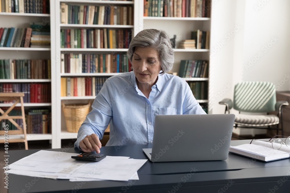 Attractive middle-aged 60s woman accountant calculating sum to pay, seated at workplace desk with laptop and bills. Female managing personal finances, control family incomes, check balance concept Attractive middle-aged 60s woman accountant calculating sum to pay, seated at workplace desk with laptop and bills. Female managing personal finances, control family incomes, check balance concept