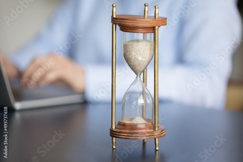 Close up view classic hourglass or sand clocks on table, business woman or office employee working on laptop seated at workplace, on background. Hurry up, deadline, productivity and efficiency concept
