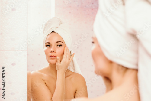 Young beautiful woman using face cream moisturizing lotion after bath. Pretty attractive girl in a towel on her head stands in front of a mirror in a home bathroom. Daily hygiene and skin care
