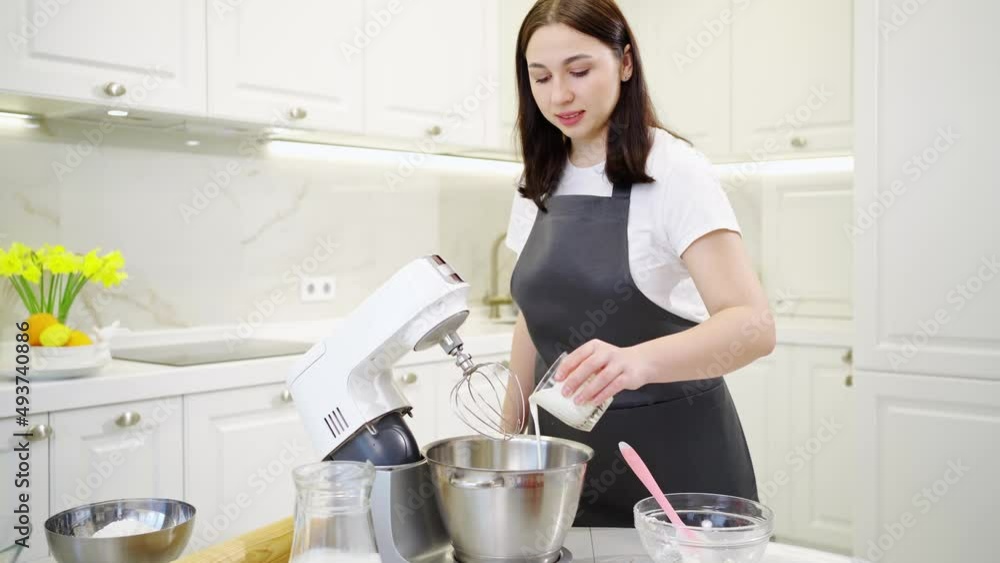 A women cook adds a milk to prepare dough or cream in a mixer bowl. A women cook adds a milk to prepare dough or cream in a mixer bowl.