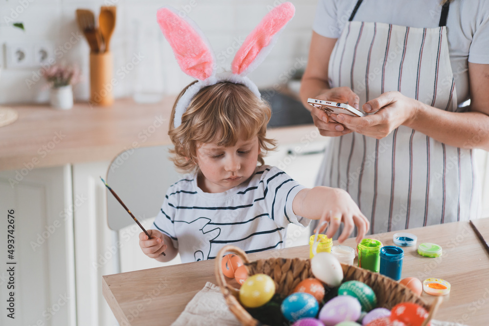 charming woman and her two-year-old son in bunny ears paint Easter eggs with paints sitting at the table on the kitchen charming woman and her two-year-old son in bunny ears paint Easter eggs with paints sitting at the table on the kitchen