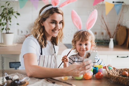 charming woman and her two-year-old son in bunny ears paint Easter eggs with paints sitting at the table on the kitchen