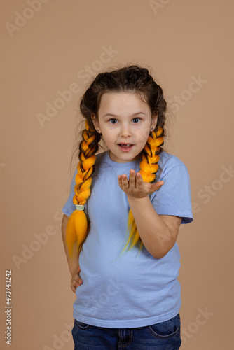 Surprised, young girl asking something showing hand looking at camera with yellow kanekalon braids on head on beige background wearing blue t-shirt and jeans.