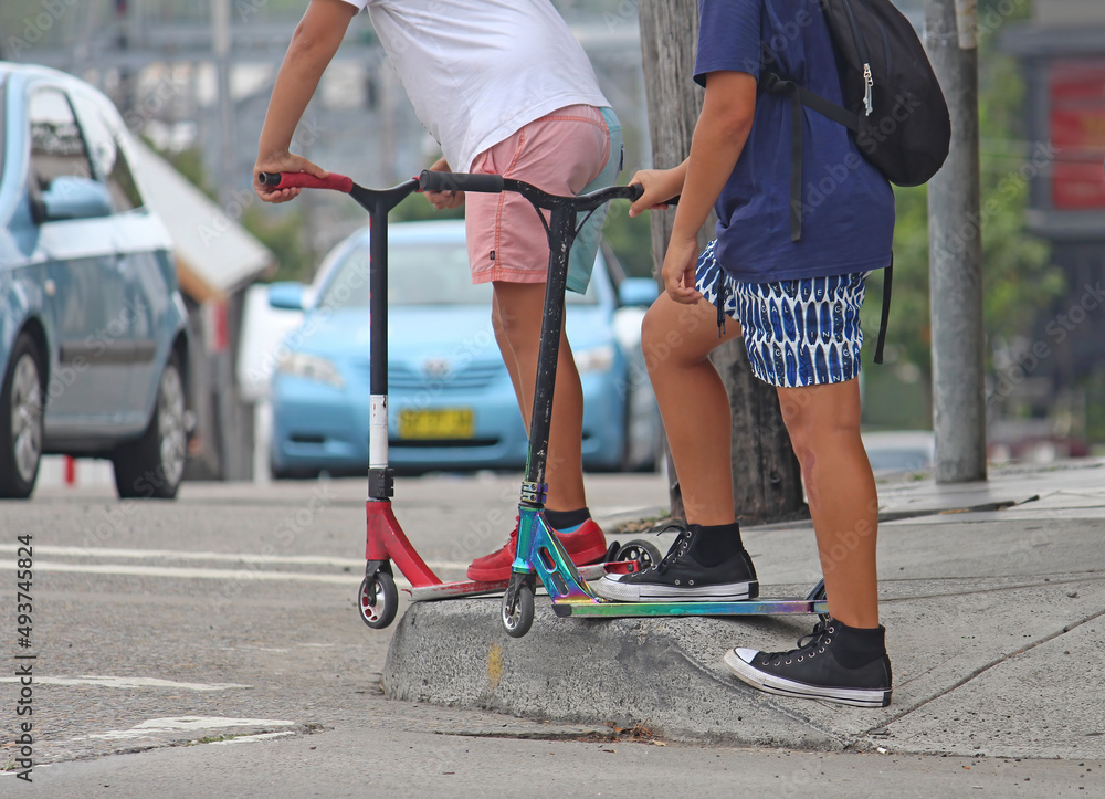 Obraz premium Two teenage boys at an intersection on scooters waiting for the traffic lights to change. Cars in the background. Generation Z.