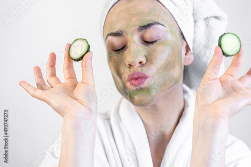 Young woman with a green face mask and fresh cucumbers in a white bathrobe.