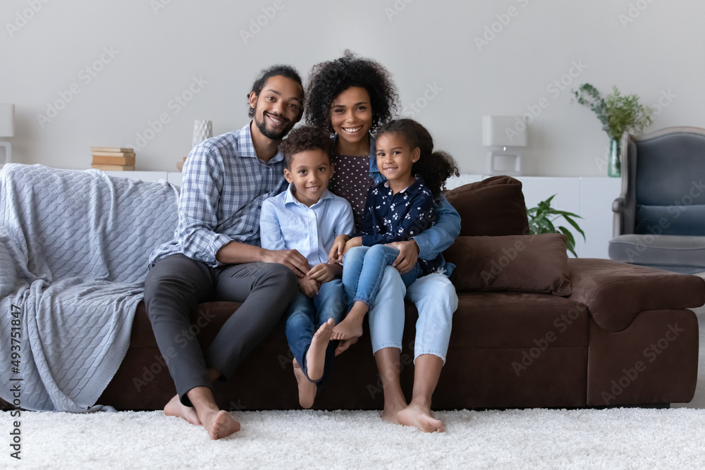 Happy perfect young Black family with two little sibling kids home portrait. Cheerful attractive couple of parents sitting on couch, holding preschool son and daughter on lap Happy perfect young Black family with two little sibling kids home portrait. Cheerful attractive couple of parents sitting on couch, holding preschool son and daughter on lap