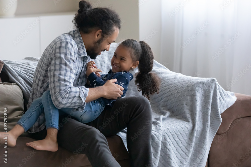 Playful happy African dad tickling cheerful little daughter kid, cuddling child on couch with love, tenderness, laughing. Young father enjoying active playtime, playing with girl at home