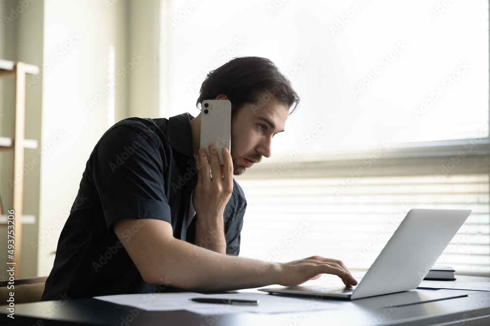 Serious focused busy manager man making telephone call to client, customer, giving consultation, using online app on laptop computer at office table, negotiating on internet project on cellphone Serious focused busy manager man making telephone call to client, customer, giving consultation, using online app on laptop computer at office table, negotiating on internet project on cellphone