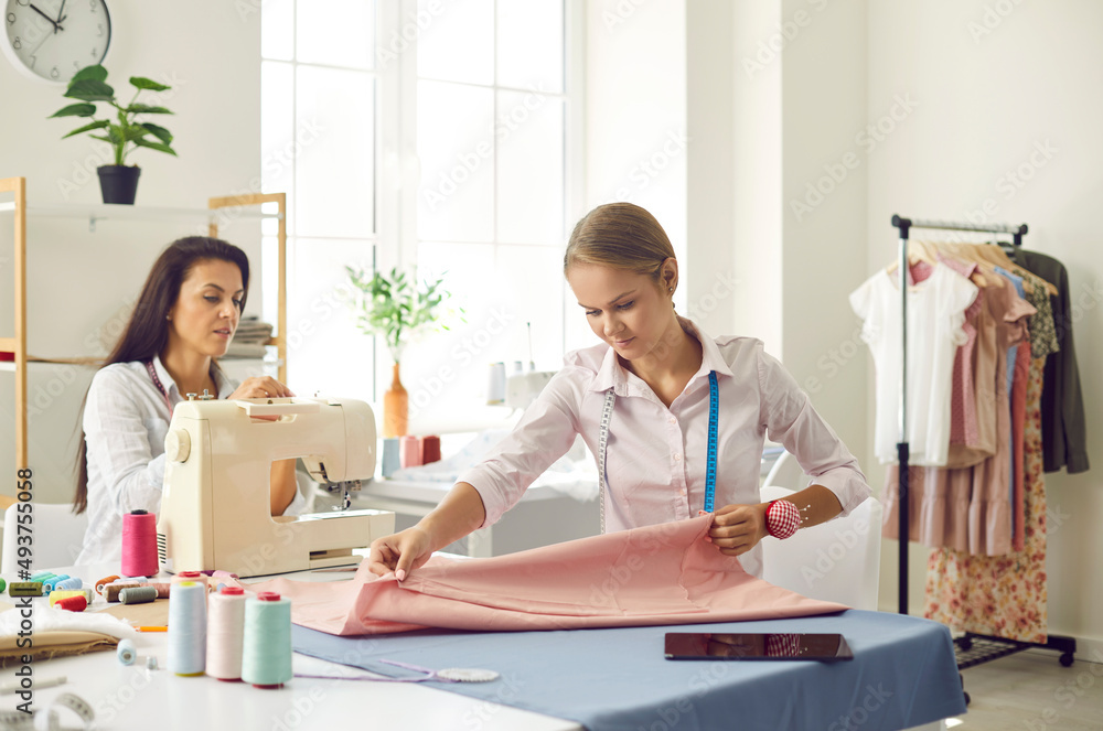 Women tailors work in own modern fashion atelier sew garment on sewing machine. Female seamstresses or dressmakers design new clothing line in workshop. Style concept. Dressmaking and manufacturing. Women tailors work in own modern fashion atelier sew garment on sewing machine. Female seamstresses or dressmakers design new clothing line in workshop. Style concept. Dressmaking and manufacturing.