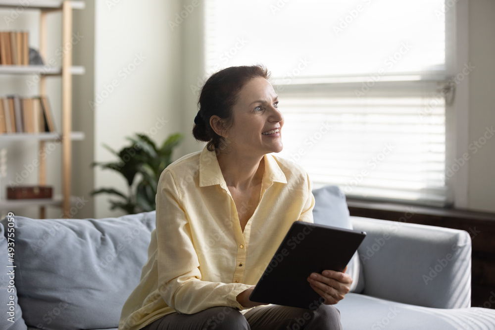 Happy thoughtful middle aged lady sitting on sofa, smiling at good thoughts, using online app on tablet, thinking over good news from internet, looking at window away. Mature woman studying online Happy thoughtful middle aged lady sitting on sofa, smiling at good thoughts, using online app on tablet, thinking over good news from internet, looking at window away. Mature woman studying online