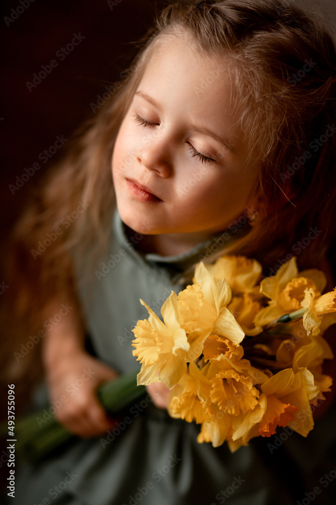 Naklejka premium beautiful spring portrait of a cute little girl with long hair and big green eyes. and a bouquet of yellow daffodils in her hand. The child enjoys the flowers and closed her eyes
