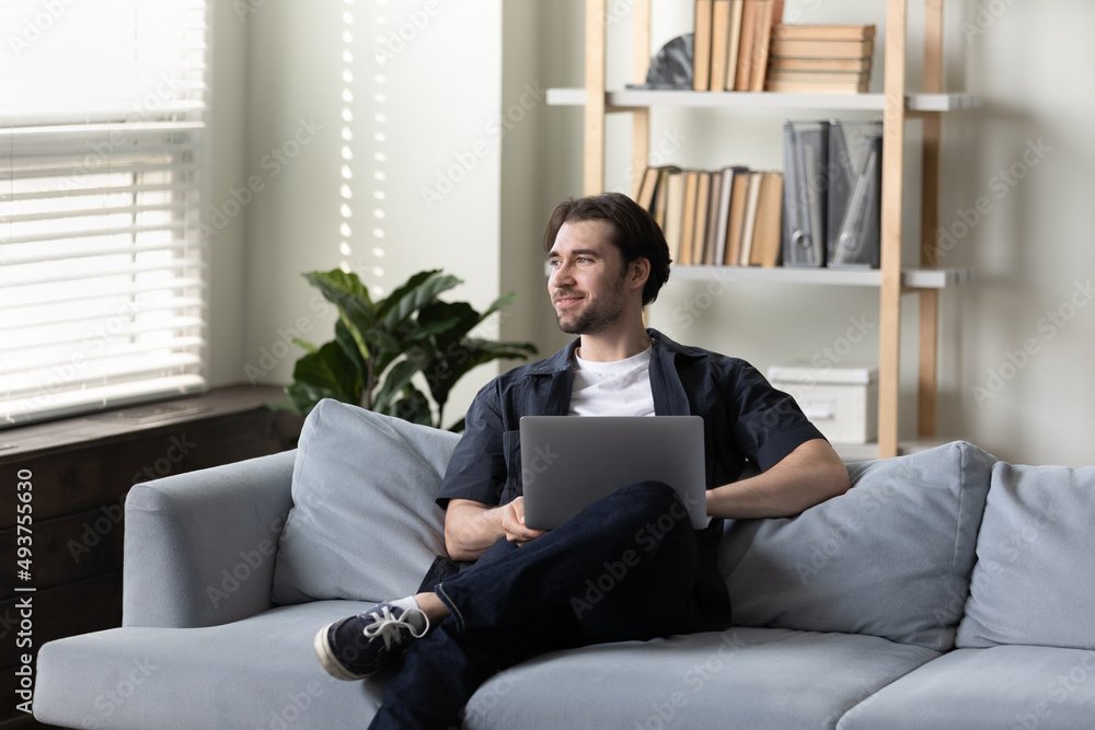 Dreamy pensive freelance man thinking over work project ideas on home office couch, using laptop, looking at window away, smiling at good thoughts, future business vision. Dreamy pensive freelance man thinking over work project ideas on home office couch, using laptop, looking at window away, smiling at good thoughts, future business vision.