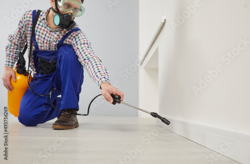 Worker of pest control service during sanitary treatment of house with insecticidal chemical sprays. Man in goggles and respirator sprays poison on floor of apartment from large spray bottle.