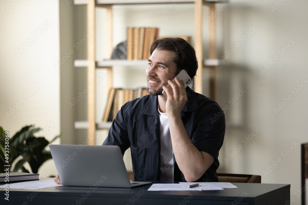 Happy handsome millennial businessman making phone call to client, consulting customer support center, speaking on cellphone at laptop computer at office workplace, looking away, smiling Happy handsome millennial businessman making phone call to client, consulting customer support center, speaking on cellphone at laptop computer at office workplace, looking away, smiling