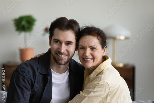 Happy mature mom embracing grown handsome son at home, looking away with toothy smile, posing for camera. Millennial guy enjoying family leisure time, meeting with mature mother. Head shot portrait