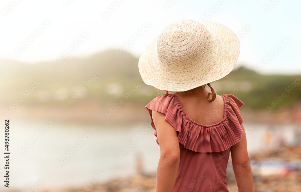 a little girl in a hat is now standing with her back to the camera on the seashore