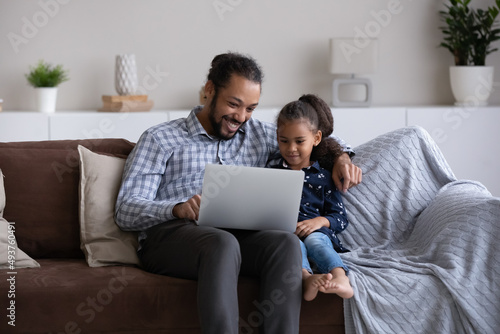 Cheerful Afro American dad and little daughter girl using learning app on laptop, watching cartoon movie together, playing educational virtual game online, reading book, resting on sofa at home