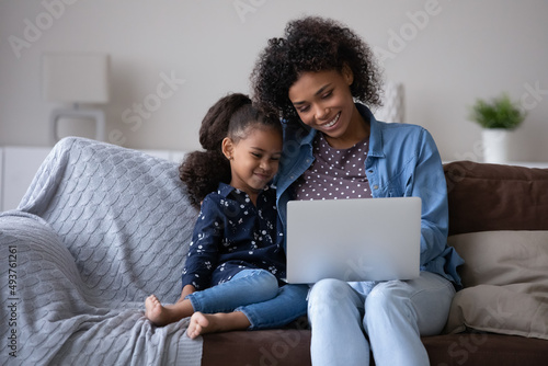 Happy sweet little daughter girl and joyful African mom reading online book on laptop, watching cartoon movie, series, sitcom, laughing, making video call, shopping together. Family communication
