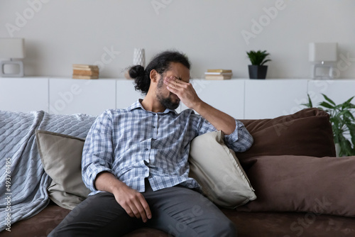 Stressed millennial African man feeling exhausted, strong headache, migraine, resting on sofa at home, touching irritable dry eyes, going through apathy, crisis. Depression, health problems concept