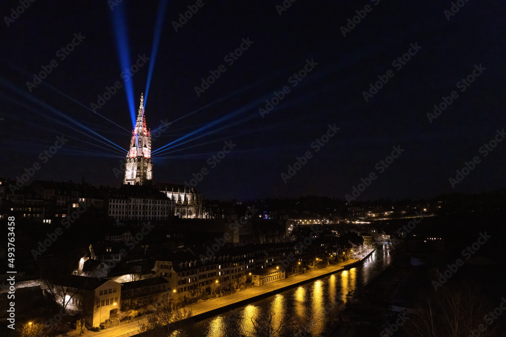 The cathedral in Bern, Switzerland, illuminated during Museumsnacht 2022, the night of museums. Several floodlights on the walls of the bell tower create rays of light in the dark sky. The Aare River