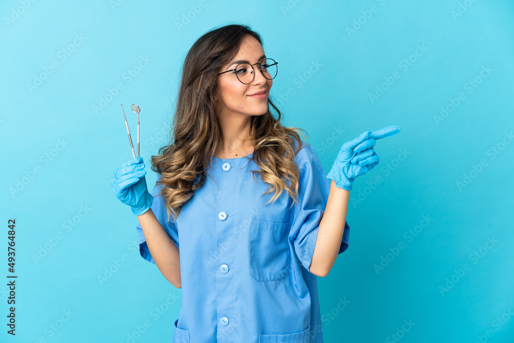 Woman dentist holding tools over isolated on blue background pointing finger to the side Woman dentist holding tools over isolated on blue background pointing finger to the side