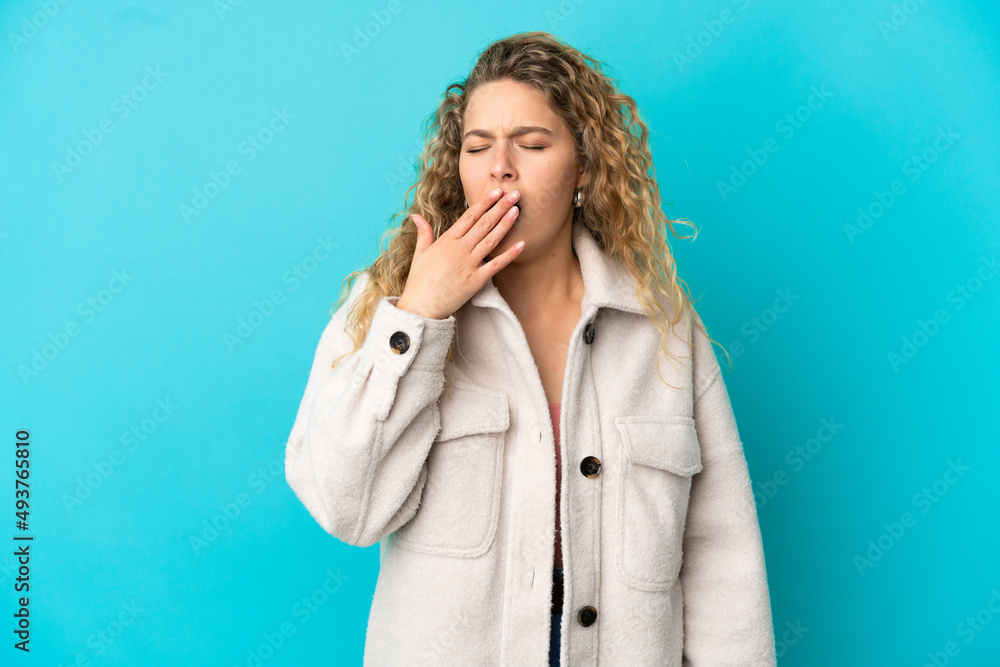 Young blonde woman isolated on blue background yawning and covering wide open mouth with hand Young blonde woman isolated on blue background yawning and covering wide open mouth with hand