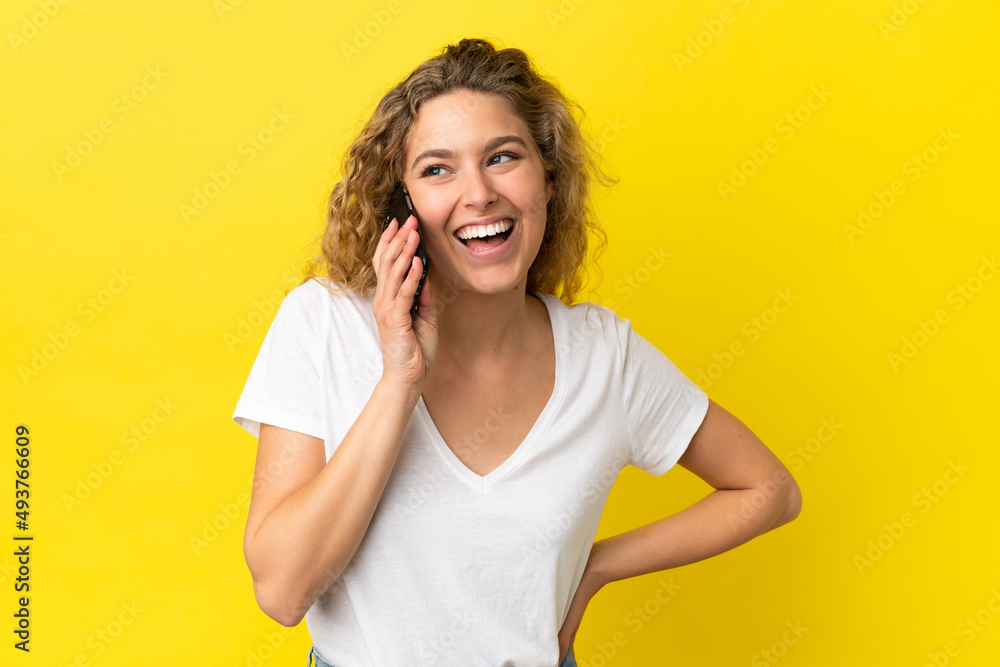 Young blonde woman using mobile phone isolated on yellow background posing with arms at hip and smiling Young blonde woman using mobile phone isolated on yellow background posing with arms at hip and smiling