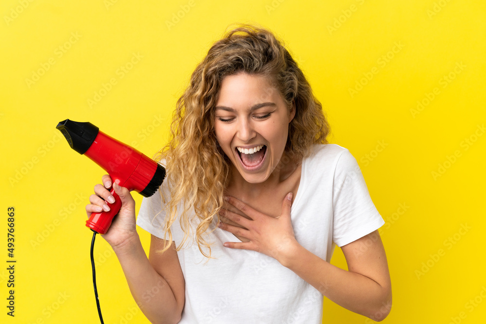 Young blonde woman holding a hairdryer isolated on yellow background smiling a lot Young blonde woman holding a hairdryer isolated on yellow background smiling a lot