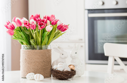 A bouquet of tulips, Easter bunnies and eggs with a golden pattern on the table. In the background is a white Scandinavian-style kitchen. Beautiful greeting card. The minimal concept.