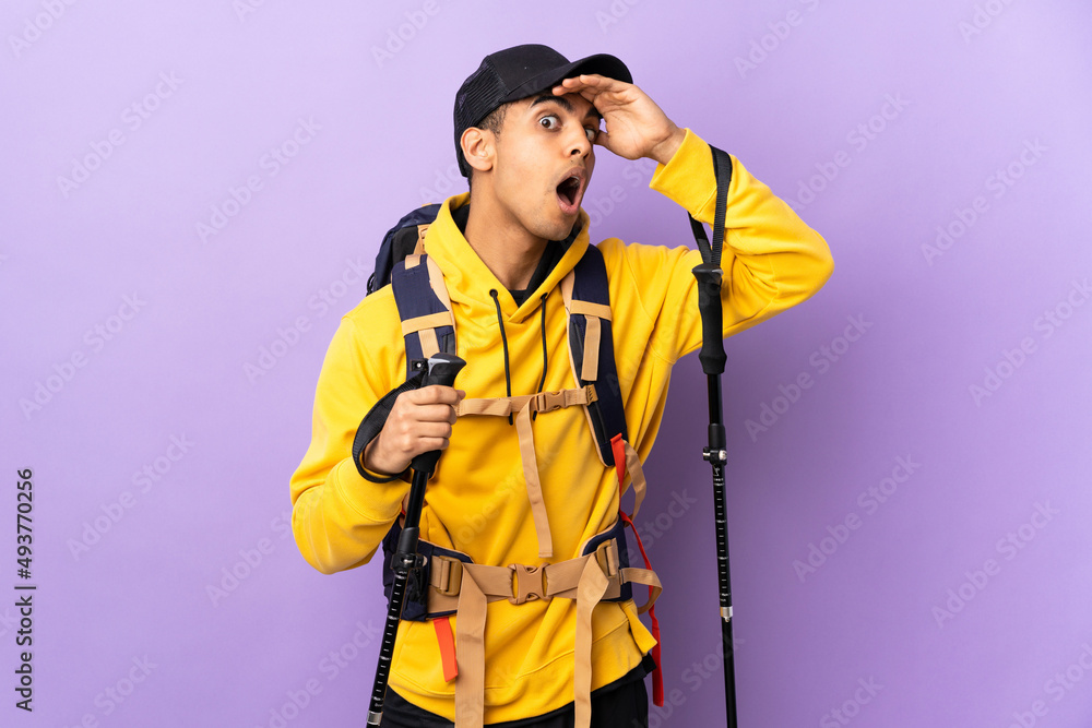 African American man with backpack and trekking poles over isolated background doing surprise gesture while looking to the side African American man with backpack and trekking poles over isolated background doing surprise gesture while looking to the side