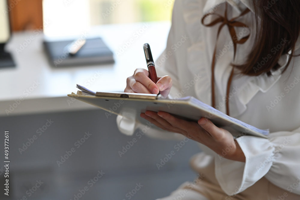 Cropped view businesswoman sitting in the office holding a report and writing something, for business and finance concept.